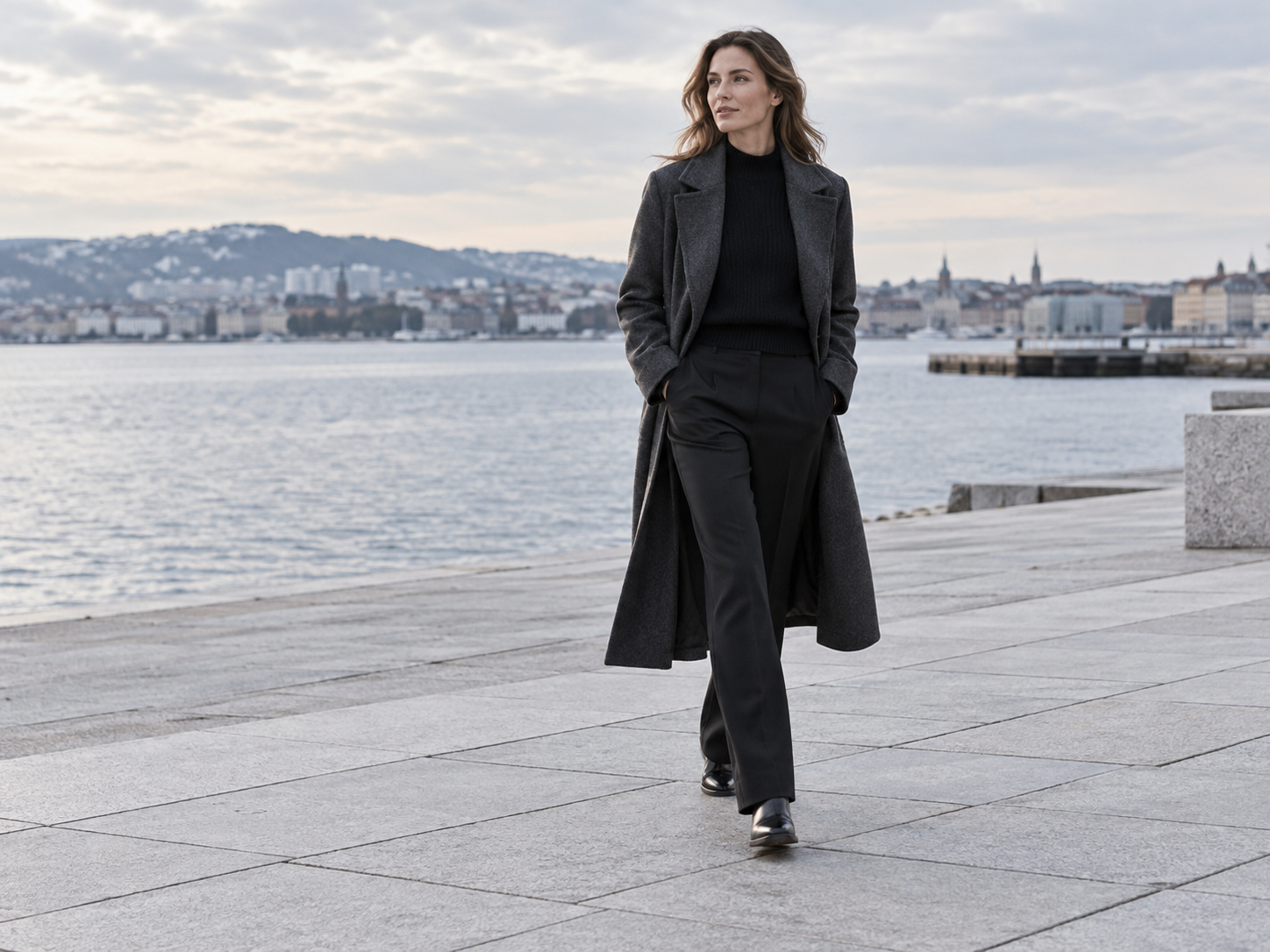 A woman styling a long charcoal coat on a waterfront plaza